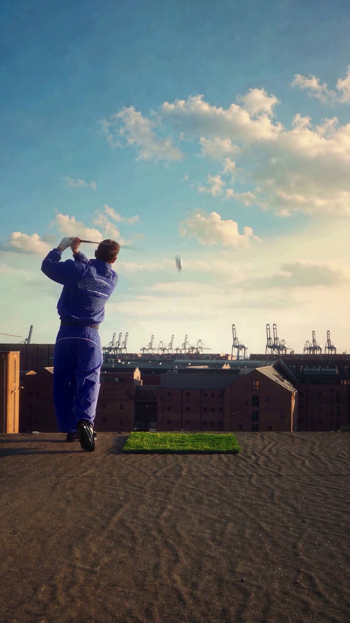 Golfer in a blue tracksuit mid-swing on a patch of turf on a rooftop above a shipping port