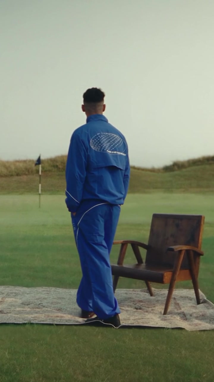 The golfer in a blue tracksuit standing beside an armchair on a rug in the middle of a fairway