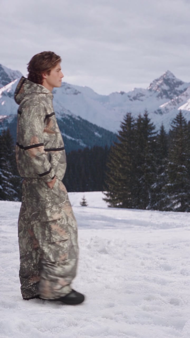 Walking across a snowfield in the leaf-camo set, mountains behind