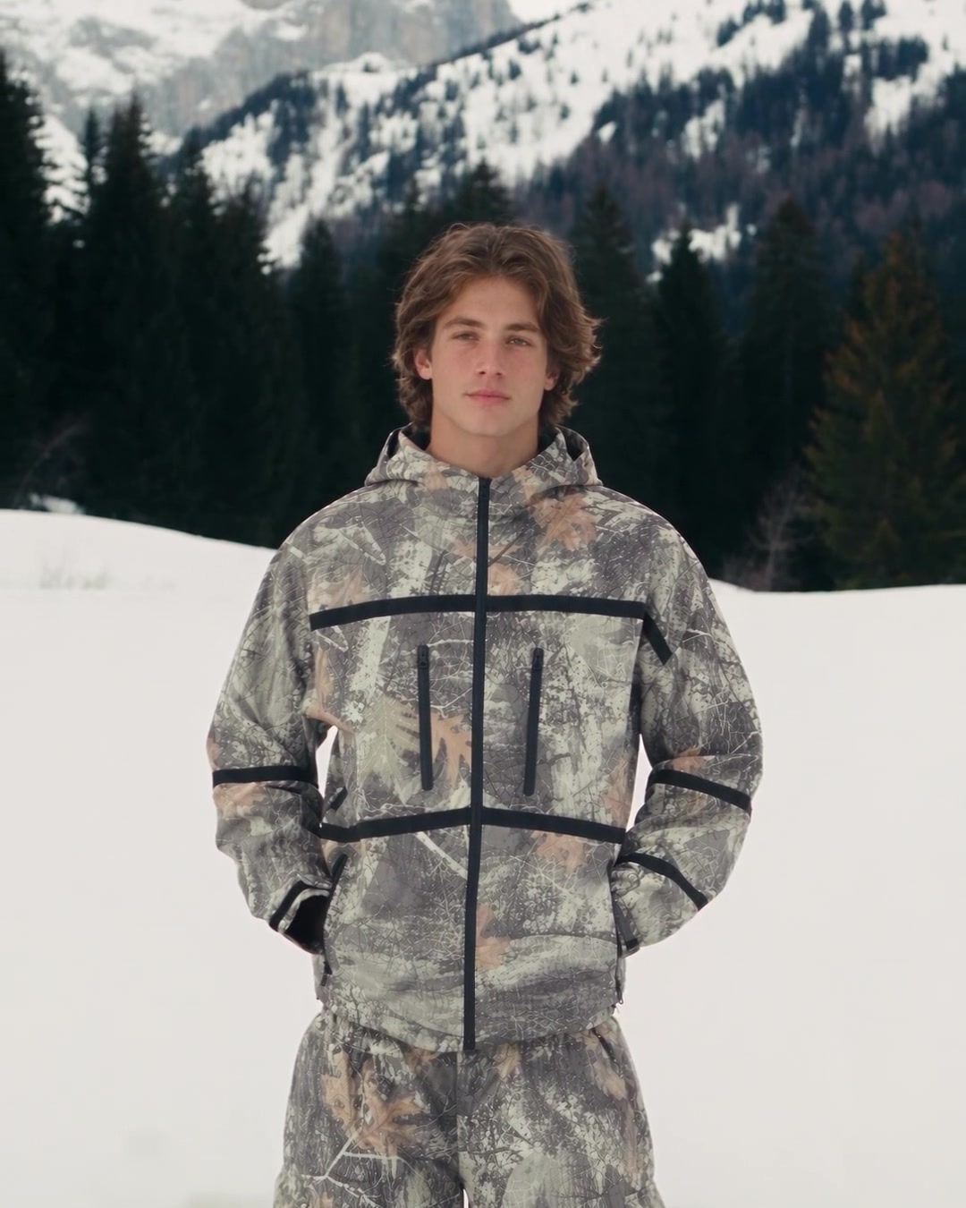 Man in a leaf-camo set standing knee-deep in snow with mountains behind